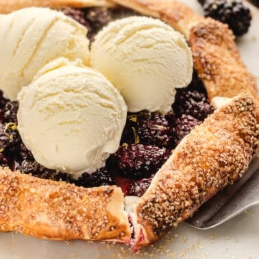 A rustic blackberry galette topped with three scoops of vanilla ice cream, served on a marble surface with a pie server.