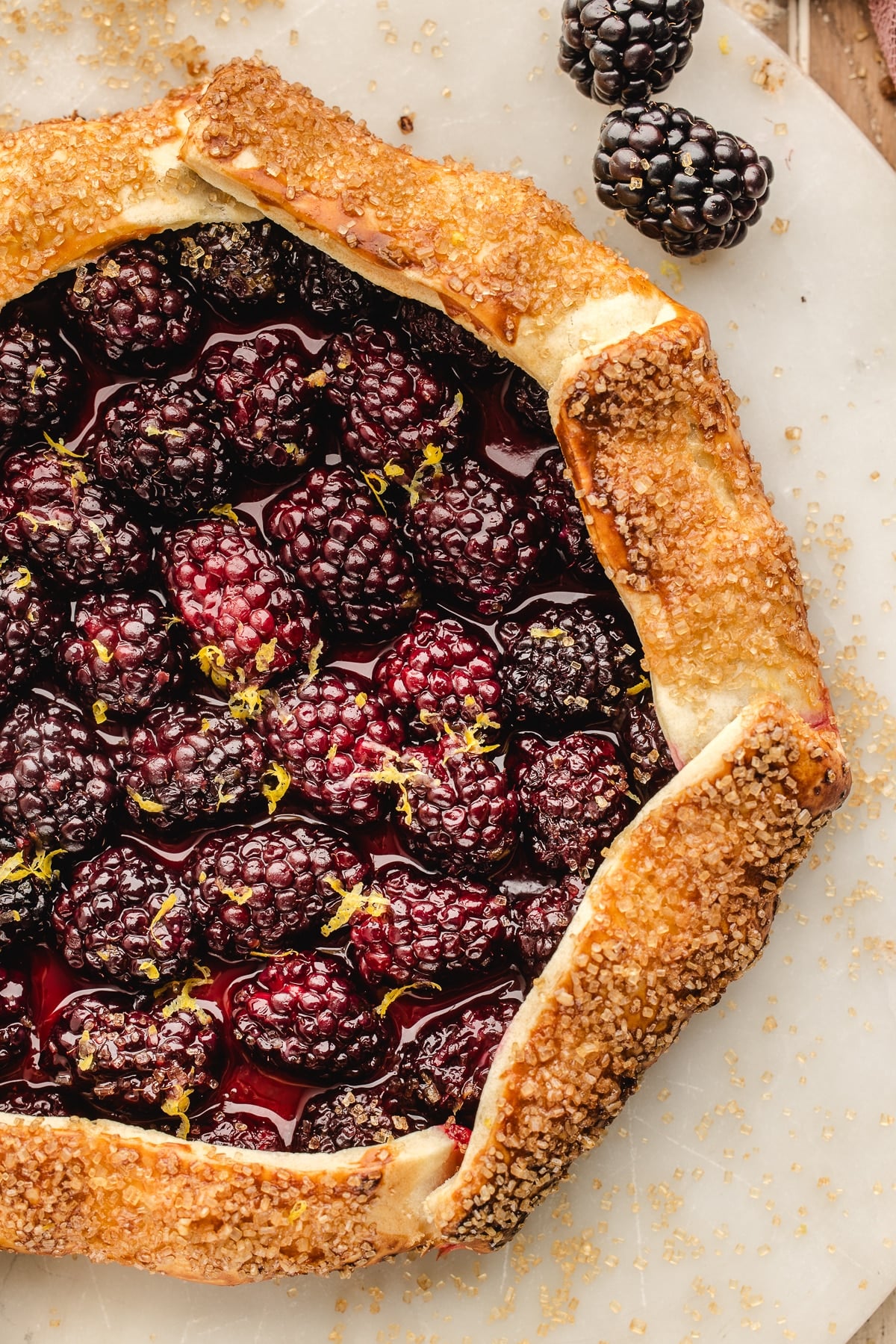 A rustic blackberry galette with a golden, sugar-coated crust is shown on a marble surface, garnished with lemon zest and fresh blackberries.