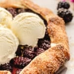 A rustic blackberry galette topped with three scoops of vanilla ice cream sits on a plate, with coarse sugar visible on the crust.