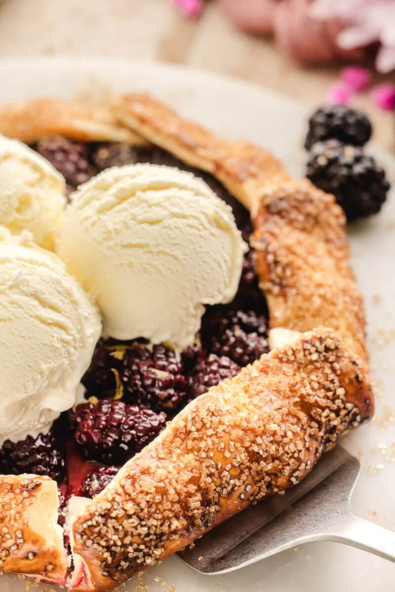A rustic blackberry galette topped with three scoops of vanilla ice cream sits on a plate, with coarse sugar visible on the crust.