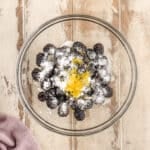 A glass bowl with blackberries, sugar, and lemon zest on a wooden surface; a folded mauve cloth sits nearby.