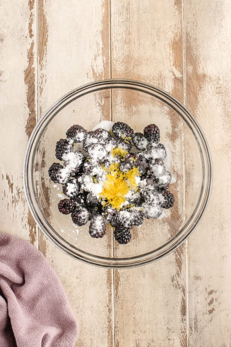 A glass bowl with blackberries, sugar, and lemon zest on a wooden surface; a folded mauve cloth sits nearby.