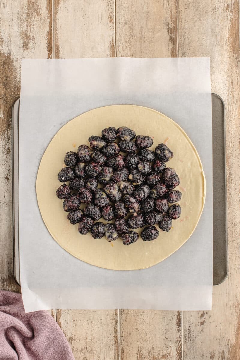 A round sheet of dough on parchment paper topped with sugared blackberries, placed on a baking tray over a wooden surface.