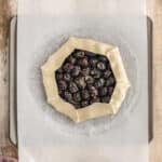 A rustic, unbaked berry galette with folded edges sits on parchment paper atop a baking sheet on a wooden surface.