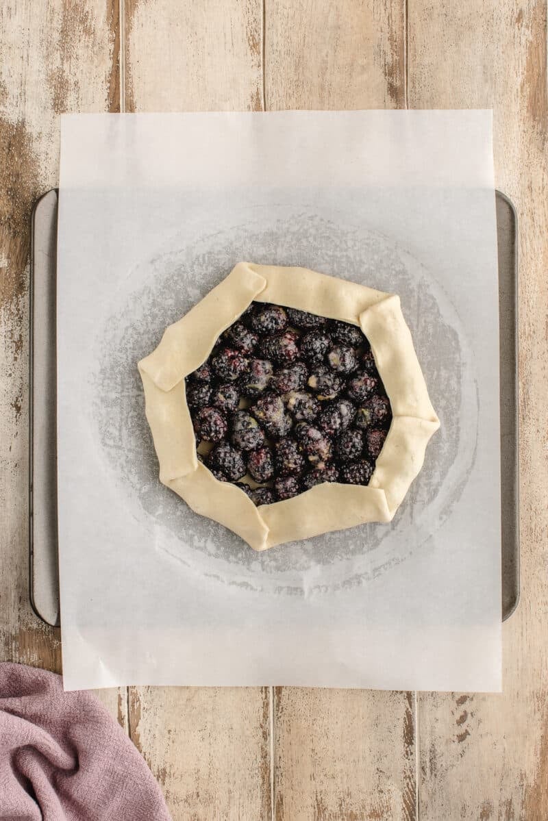 A rustic, unbaked berry galette with folded edges sits on parchment paper atop a baking sheet on a wooden surface.