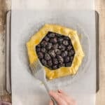 A hand brushes egg wash onto the crust of an unbaked blackberry galette on parchment paper atop a baking sheet.