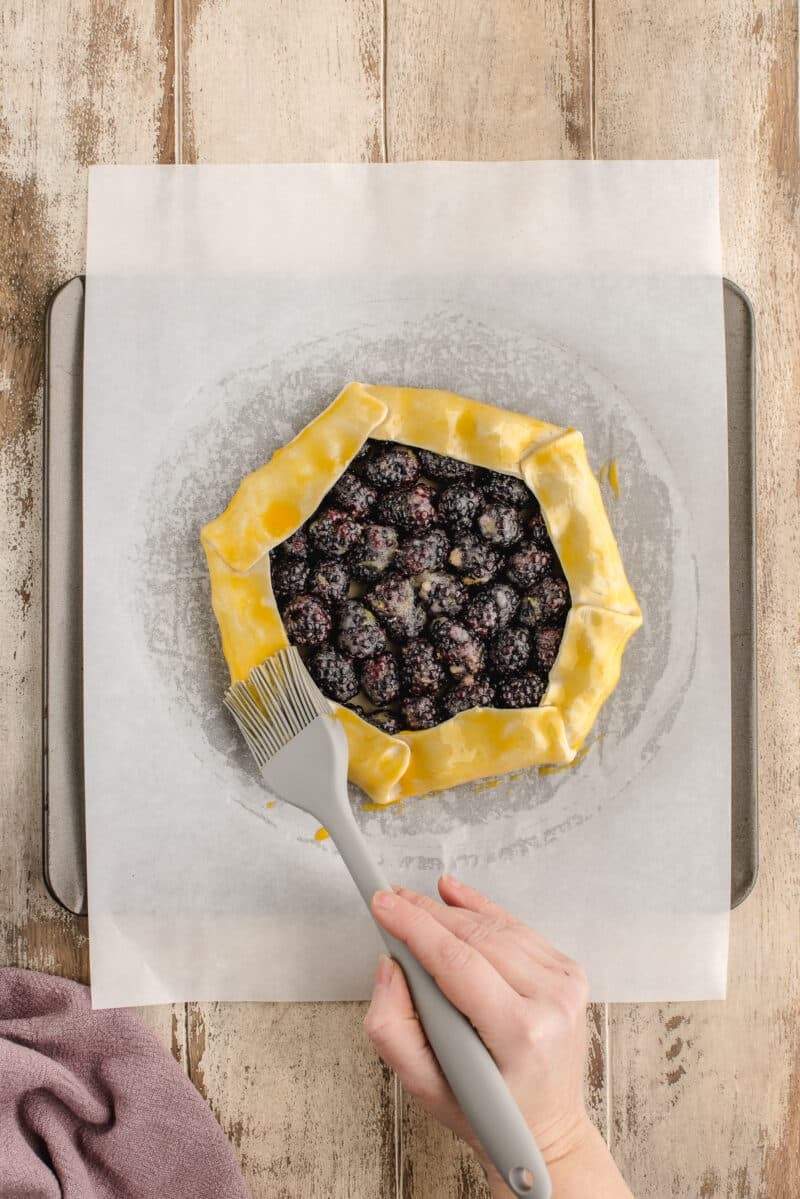 A hand brushes egg wash onto the crust of an unbaked blackberry galette on parchment paper atop a baking sheet.