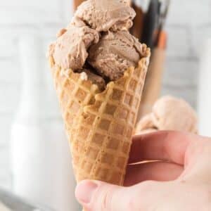 A hand is holding a waffle cone filled with several scoops of light brown ice cream. A blurred background shows a milk bottle and utensils.