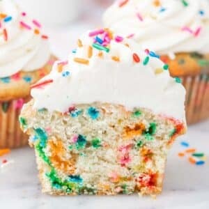 A sliced funfetti cupcake with colorful sprinkles inside and white frosting topped with more sprinkles.