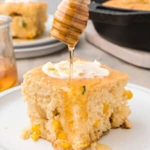 A square piece of cornbread with visible corn kernels, topped with butter, is being drizzled with honey from a dipper on a white plate.