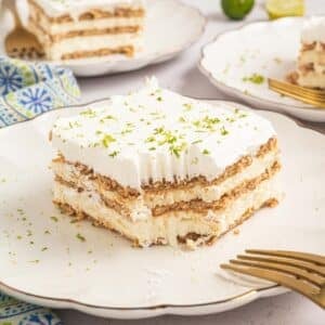 A slice of key lime icebox cake with cream and graham crackers, topped with lime zest, sits on a white plate with a fork beside it.