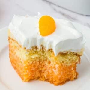 A slice of orange poke cake with white whipped topping and a mandarin orange segment on a white plate.