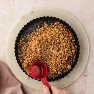 A red measuring cup pours liquid over a crumb crust in a tart pan.