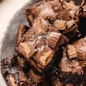 A close-up of chocolate brownies topped with chopped peanut butter cups and a sprinkle of sea salt, stacked on a plate.