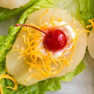 A pear half on a lettuce leaf topped with a dollop of mayonnaise, shredded cheddar cheese, and a maraschino cherry.