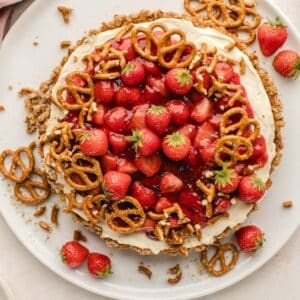 A round strawberry pretzel tart with a crust, cream cheese filling, and topped with sliced strawberries, strawberry glaze, and mini pretzels.