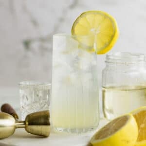 A tall glass of Vodka Collins garnished with a lemon slice sits on a surface next to a jar, a cocktail jigger, and a halved lemon.