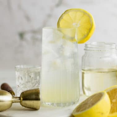 A tall glass of Vodka Collins garnished with a lemon slice sits on a surface next to a jar, a cocktail jigger, and a halved lemon.