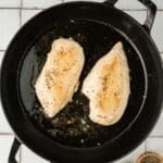 Two seasoned chicken breasts are cooking in a black cast iron skillet, with small bowls of salt and pepper beside the pan on a tiled surface.
