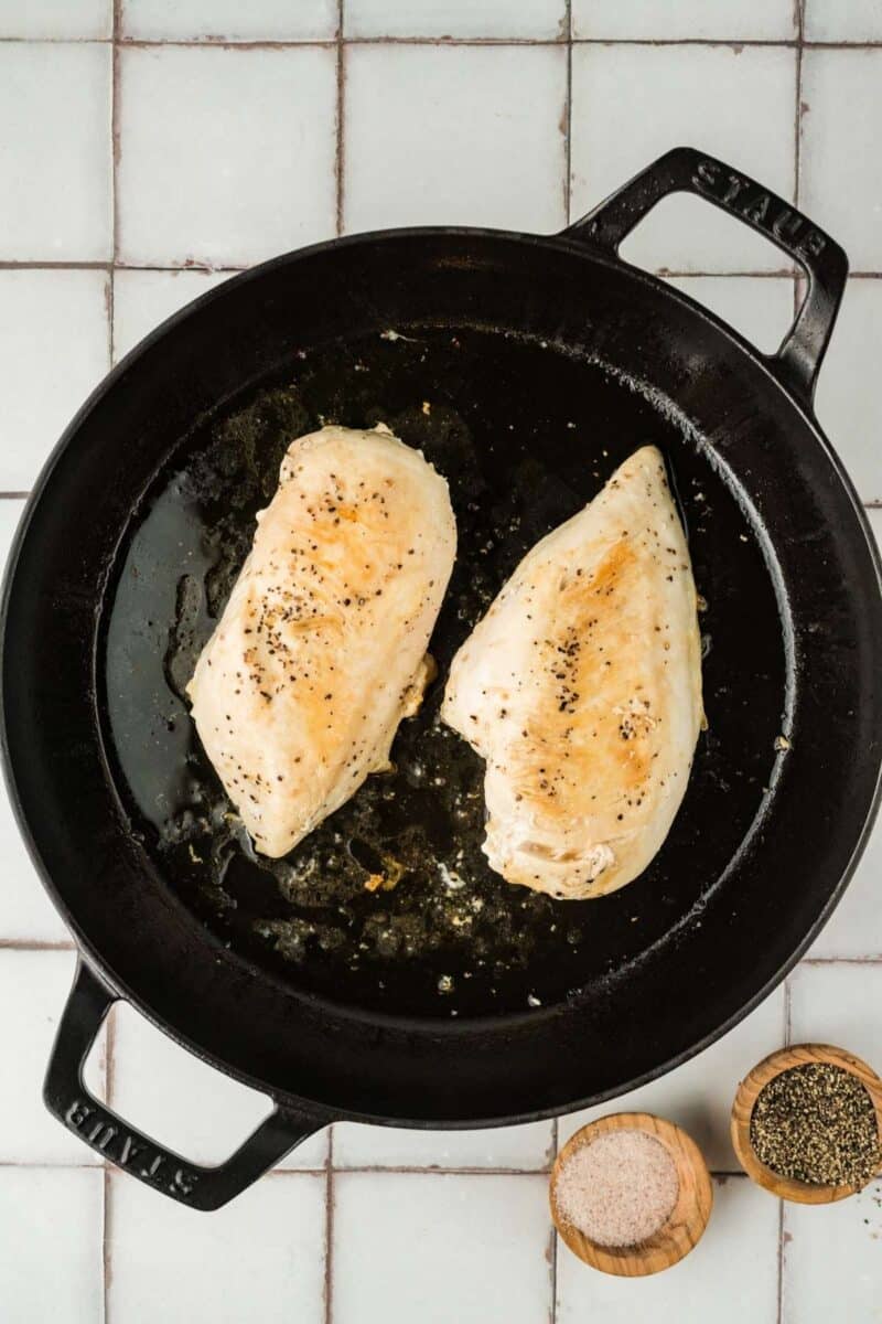 Two seasoned chicken breasts are cooking in a black cast iron skillet, with small bowls of salt and pepper beside the pan on a tiled surface.