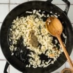 Chopped onions and seasonings cooking in a black cast iron pan with a wooden spoon, on a tiled surface next to small bowls of salt and pepper.