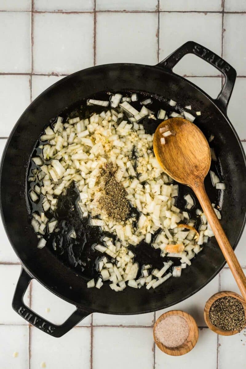 Chopped onions and seasonings cooking in a black cast iron pan with a wooden spoon, on a tiled surface next to small bowls of salt and pepper.