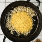A black skillet with sautéed chopped onions and dry orzo pasta, next to small wooden bowls of salt and pepper on a tiled surface.