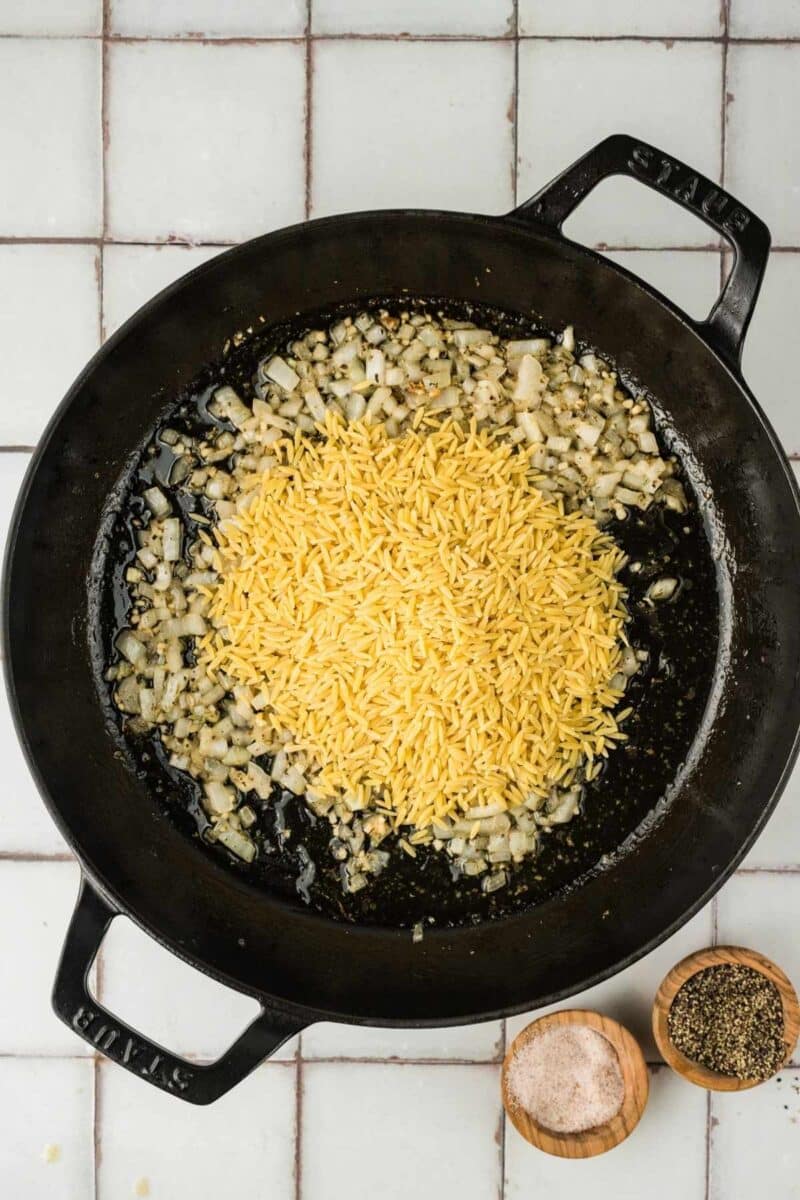 A black skillet with sautéed chopped onions and dry orzo pasta, next to small wooden bowls of salt and pepper on a tiled surface.