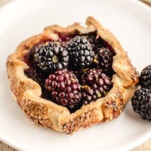 A small rustic blackberry tart with a golden, sugared crust sits on a white plate, topped with fresh blackberries.