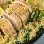 Sliced cooked chicken breast served on creamy orzo with spinach, asparagus, and lemon slices in a black pan.