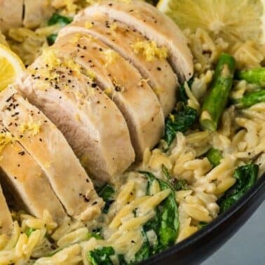 Sliced cooked chicken breast served on creamy orzo with spinach, asparagus, and lemon slices in a black pan.