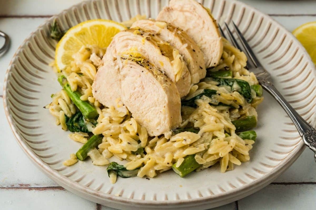 Sliced cooked chicken breast served on a bed of orzo pasta with spinach and asparagus, garnished with a lemon wedge on a beige plate.