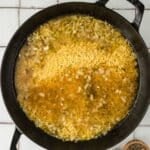 A black pot with uncooked orzo, chopped onions, and broth on a tiled surface, with small bowls of salt and pepper beside it.