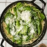 A skillet filled with creamy pasta topped with fresh spinach leaves and grated cheese, placed on a tiled surface next to small bowls of salt and pepper.