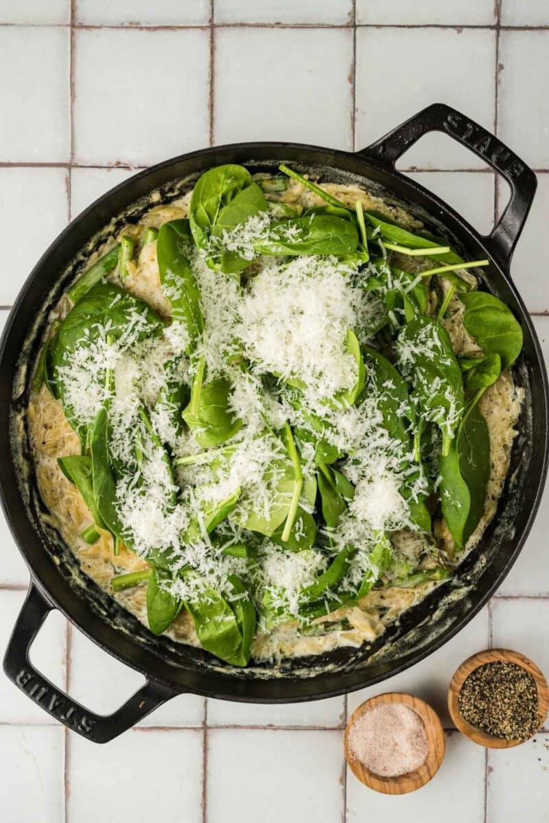 A skillet filled with creamy pasta topped with fresh spinach leaves and grated cheese, placed on a tiled surface next to small bowls of salt and pepper.