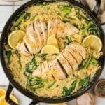 A skillet filled with creamy orzo, spinach, asparagus, and sliced cooked chicken breast, garnished with lemon wedges. Plates, utensils, and ingredients are arranged nearby.