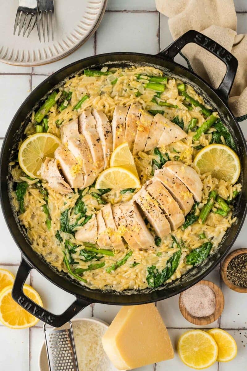 A skillet filled with creamy orzo, spinach, asparagus, and sliced cooked chicken breast, garnished with lemon wedges. Plates, utensils, and ingredients are arranged nearby.