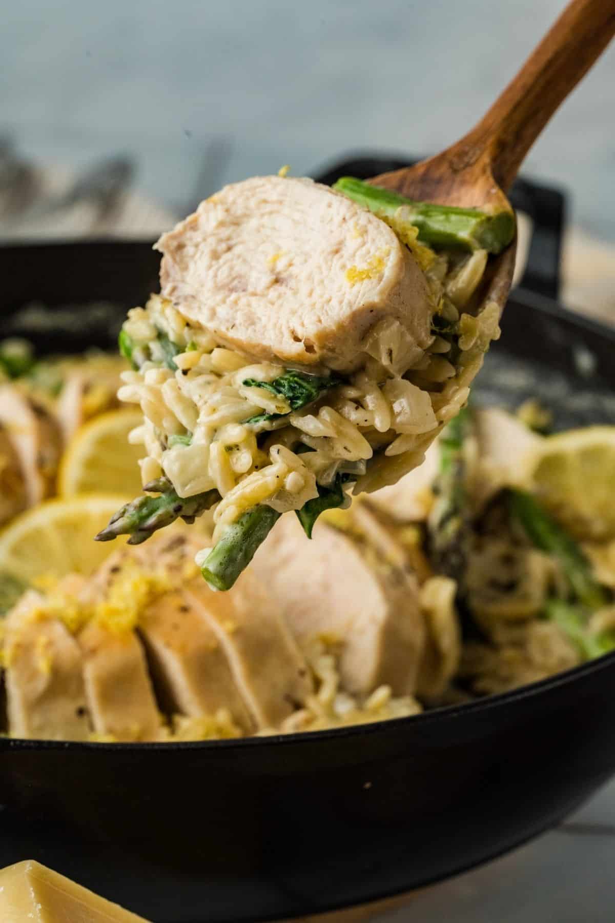 A wooden spoon holds a serving of creamy pasta with asparagus and a slice of cooked chicken breast over a pan with more pasta, chicken, and lemon slices.