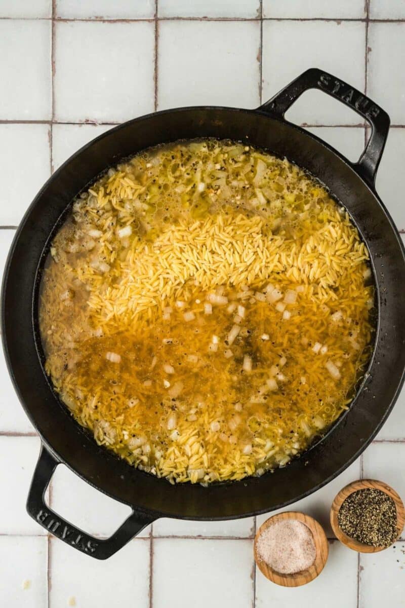 A black pot with uncooked orzo, chopped onions, and broth on a tiled surface, with small bowls of salt and pepper beside it.