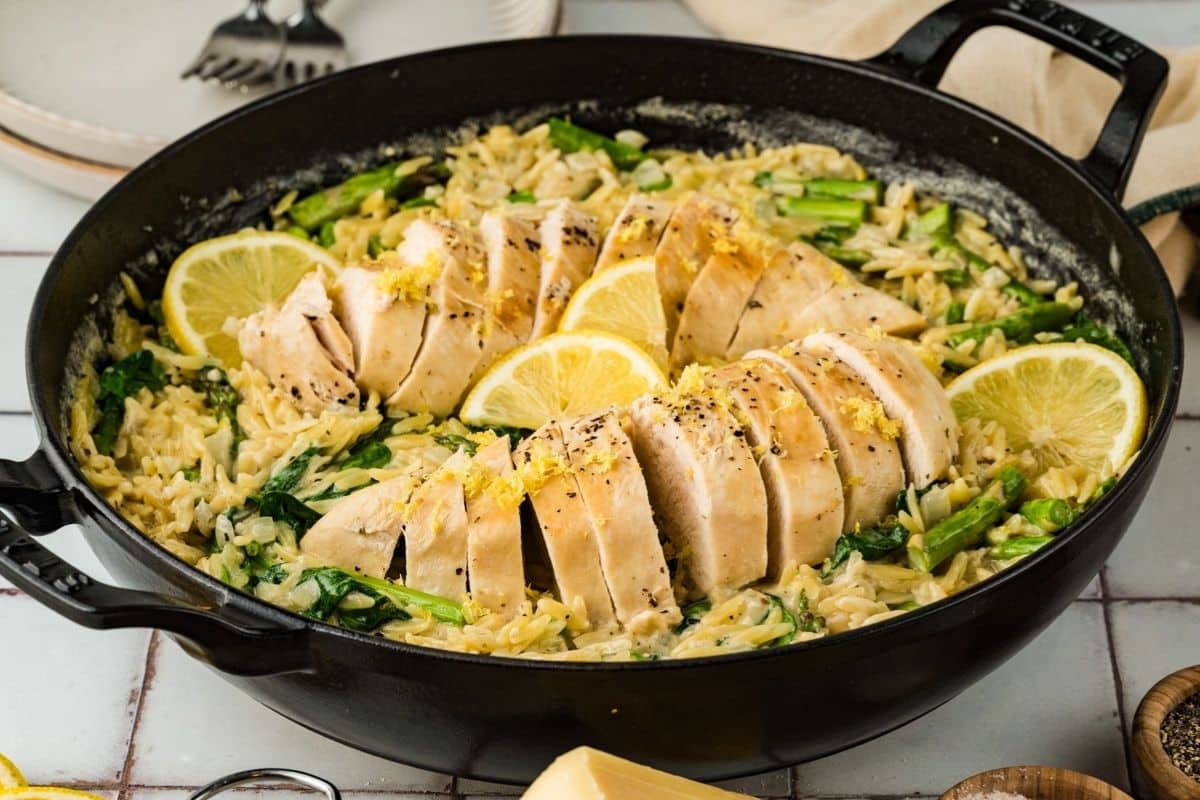 Sliced cooked chicken breast on top of creamy orzo with spinach and asparagus, garnished with lemon slices, served in a black cast iron pan.