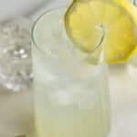 A clear glass filled with a light-colored drink, ice cubes, and garnished with a lemon slice on the rim. Other glasses and a stirring stick are in the background.