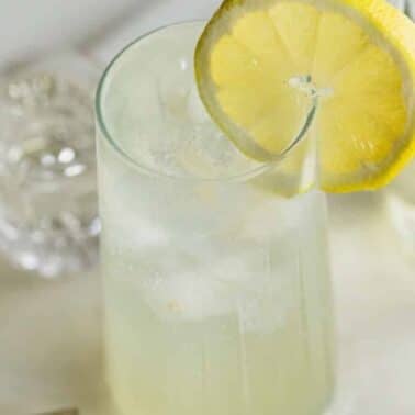 A clear glass filled with a light-colored drink, ice cubes, and garnished with a lemon slice on the rim. Other glasses and a stirring stick are in the background.