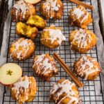 Twelve apple fritters with white icing drizzle are arranged on a cooling rack, with apple slices and cinnamon sticks placed nearby.