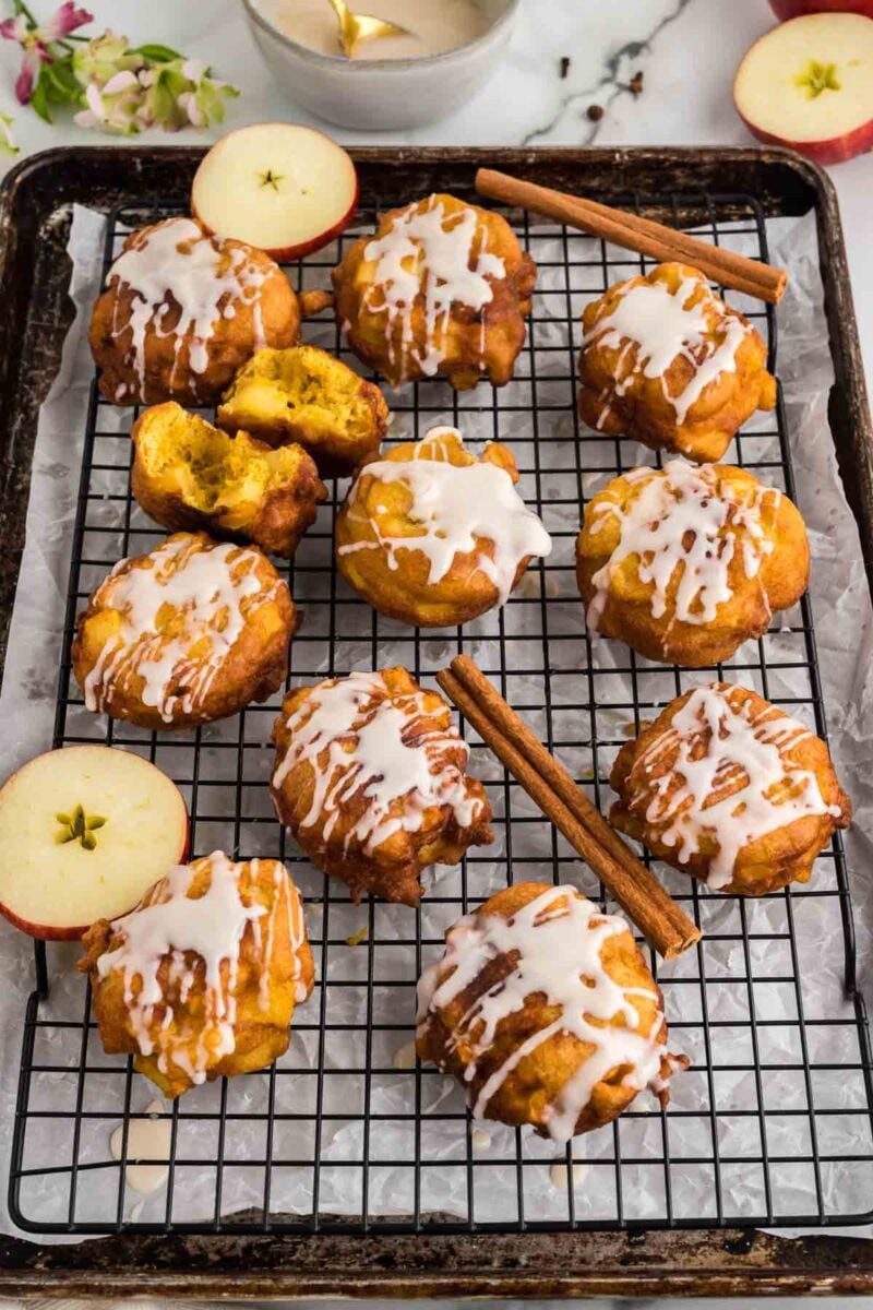 Twelve apple fritters with white icing drizzle are arranged on a cooling rack, with apple slices and cinnamon sticks placed nearby.