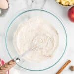 A glass bowl of flour mixture with a whisk, surrounded by eggs, vanilla, milk, diced apples, a peach, cinnamon sticks, and a striped towel on a marble surface.
