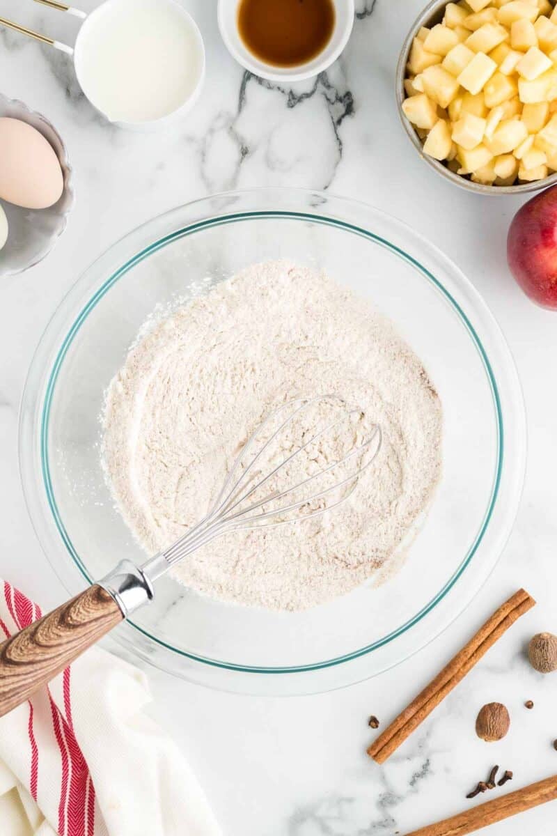 A glass bowl of flour mixture with a whisk, surrounded by eggs, vanilla, milk, diced apples, a peach, cinnamon sticks, and a striped towel on a marble surface.