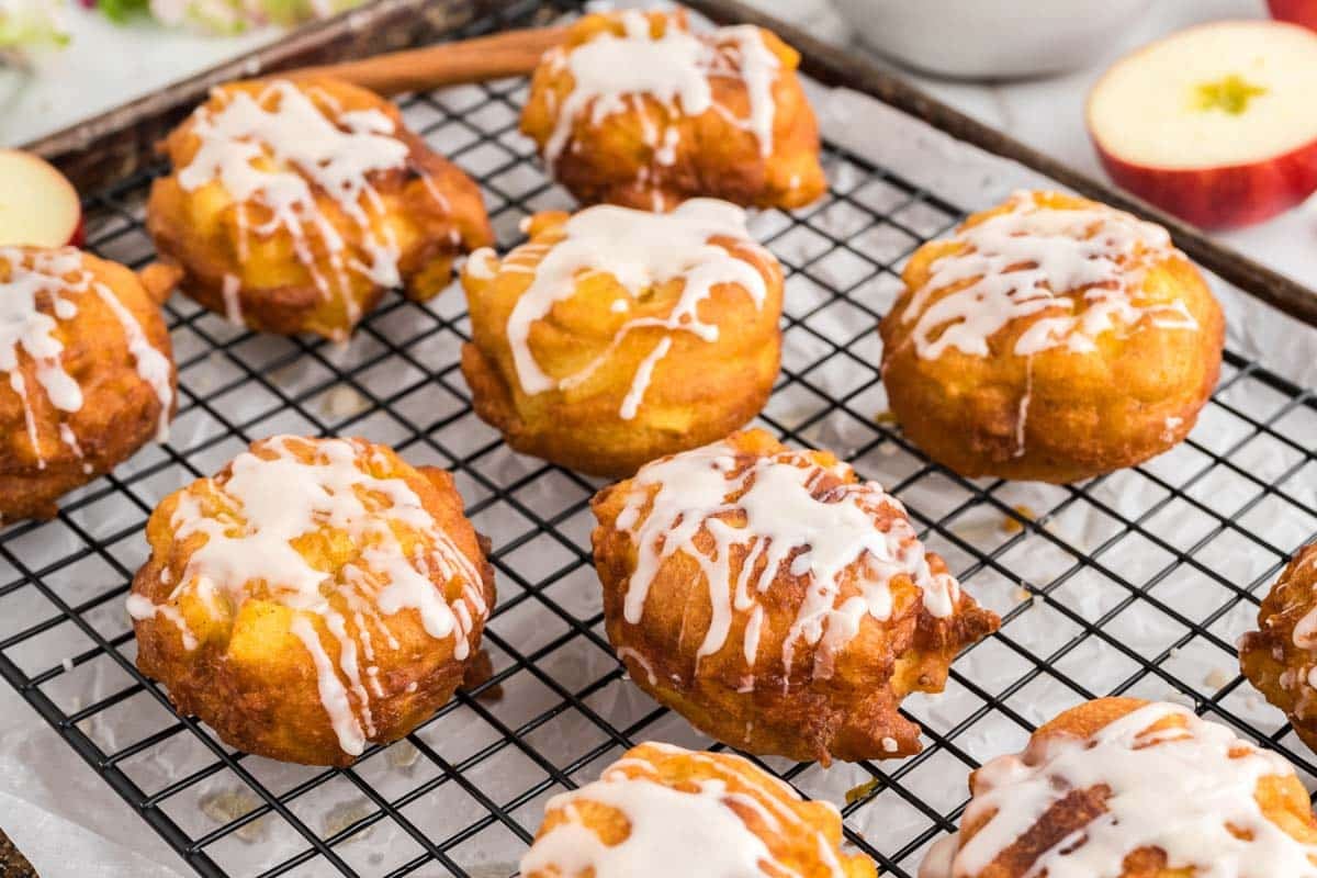 A cooling rack holds several glazed apple fritters with icing drizzled on top; a sliced apple is visible in the background.