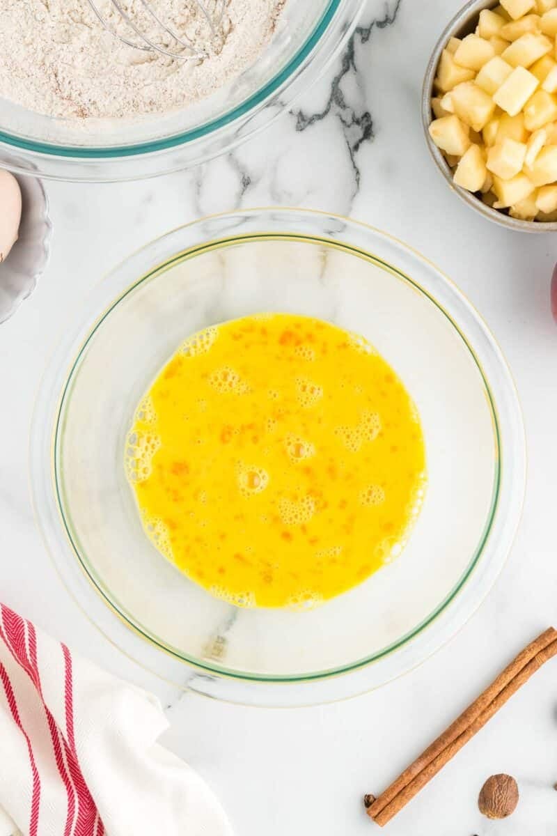 A clear glass bowl filled with beaten eggs sits on a marble countertop, surrounded by chopped apples, a bowl of flour, a whisk, a cinnamon stick, and a red-striped towel.