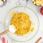 A glass bowl of yellow cake batter with a spatula, surrounded by chopped apples, eggshells, powdered sugar, a red apple, cinnamon sticks, and spices on a marble countertop.
