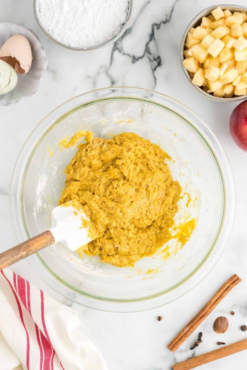 A glass bowl of yellow cake batter with a spatula, surrounded by chopped apples, eggshells, powdered sugar, a red apple, cinnamon sticks, and spices on a marble countertop.